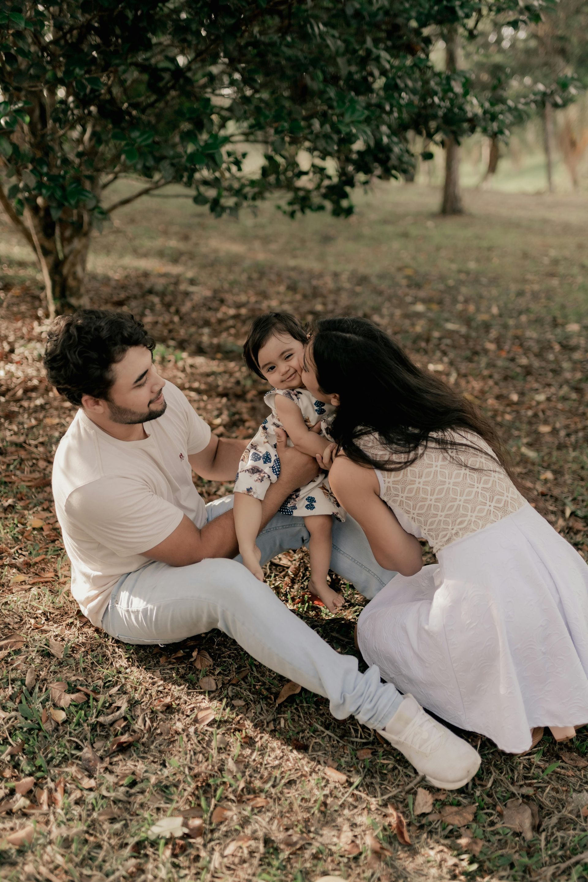 A family of three sitting on dry grass under a tree, with the parents embracing their child.