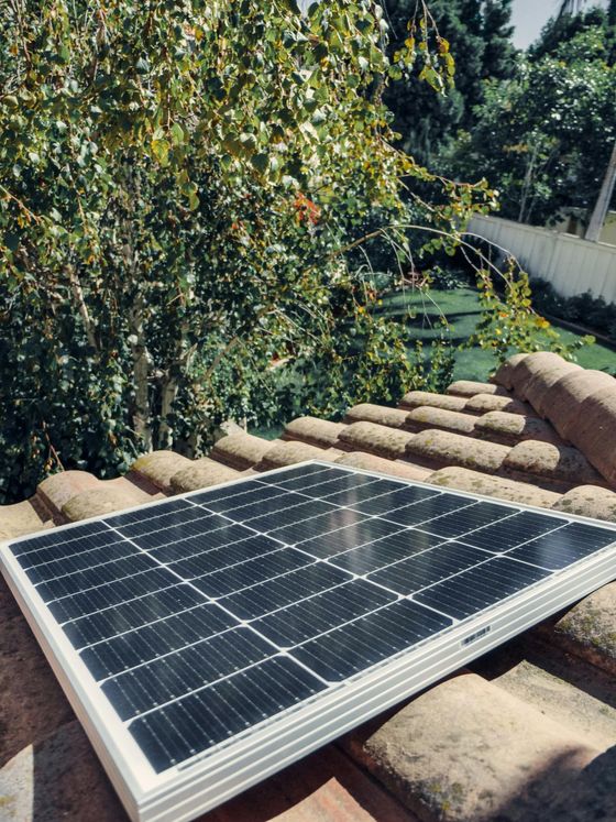 Solar panel on a terracotta tile roof with a blurred green tree and white fence in the background.
