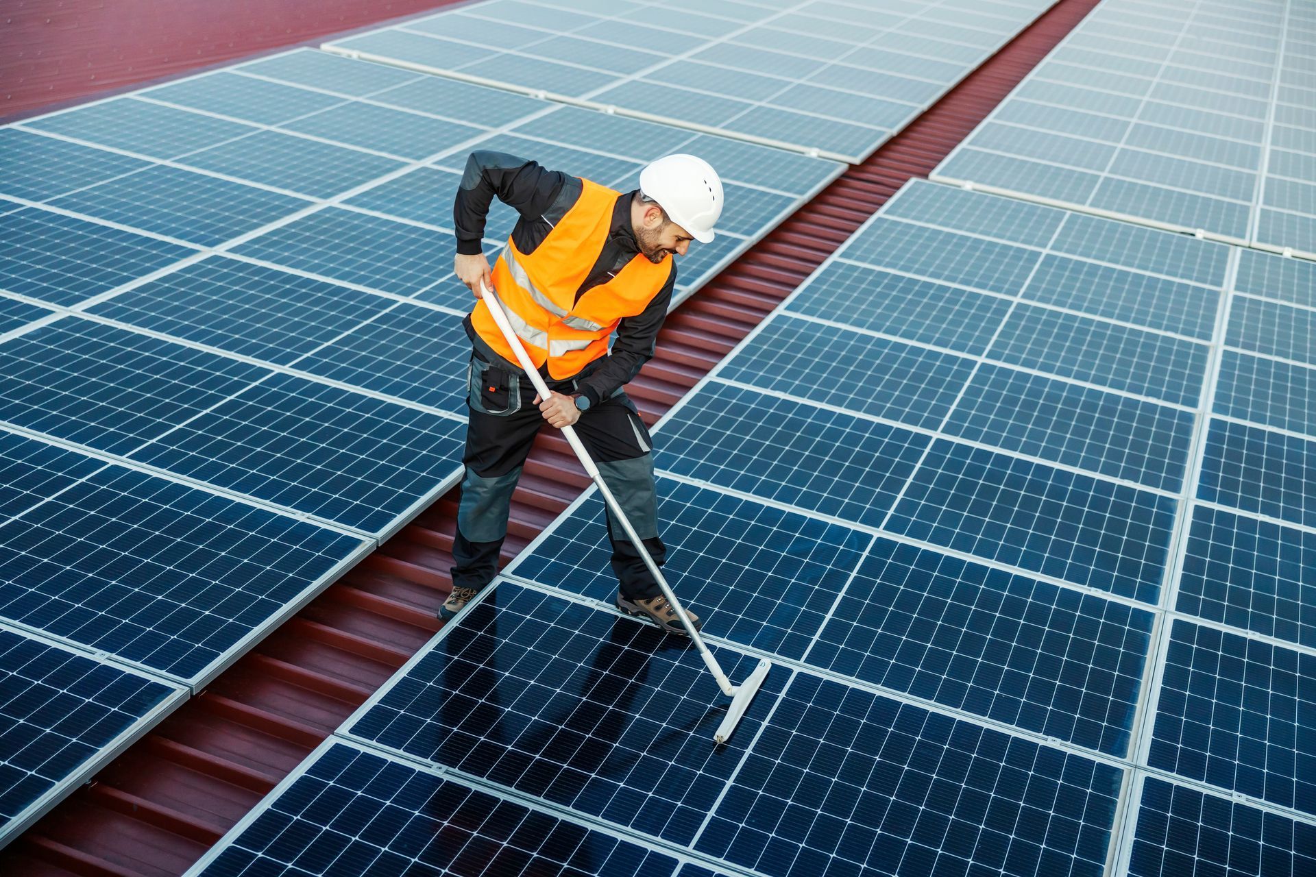 Worker in safety vest and hard hat cleaning solar panels on a rooftop with a long-handled squeegee.