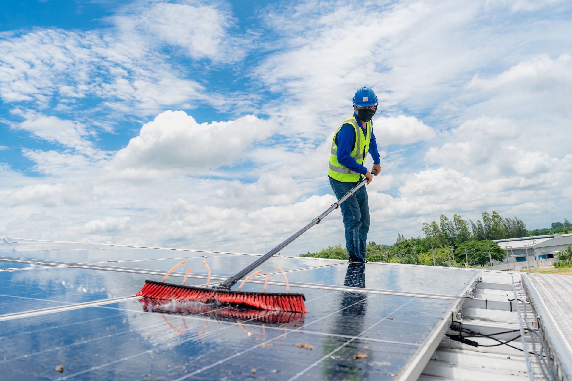 Person in safety gear cleaning solar panels on a roof with a long-handled brush under a blue sky.