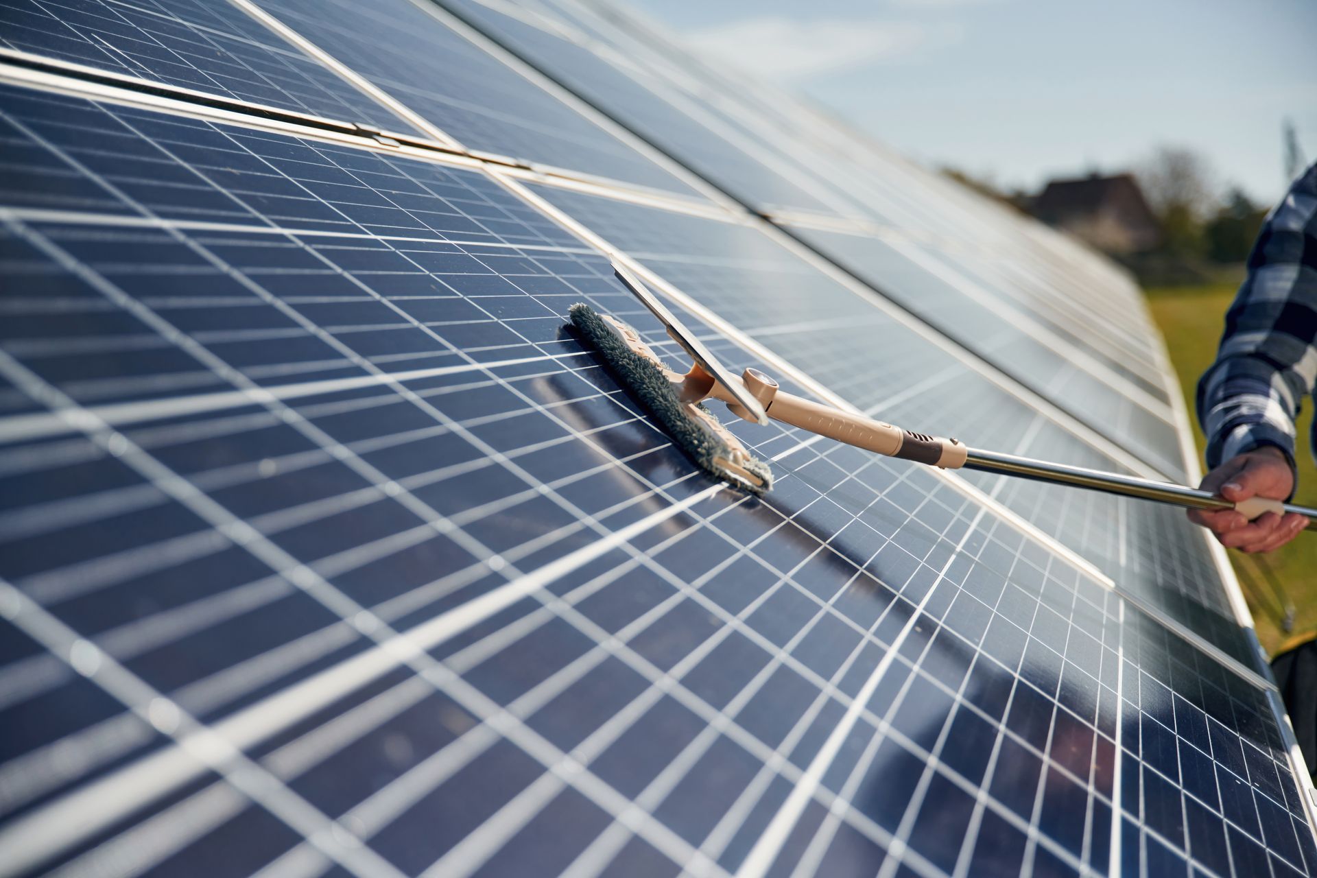 Person cleaning solar panels with a squeegee. The panels are installed outdoors in a field, reflecting the blue sky.