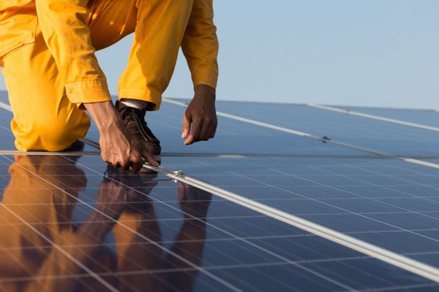 Person in yellow jumpsuit working on solar panels on a rooftop under a clear sky.