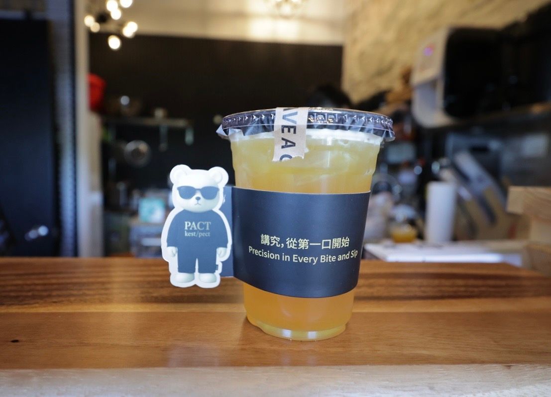 Iced drink with blue sleeve beside a small bear figurine on a wooden counter in a cafe