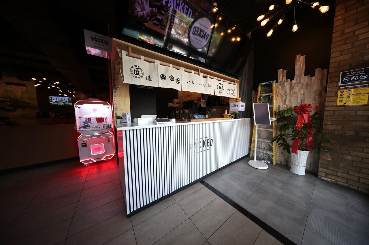 Small striped-front bar kiosk with menu sign, red-lit vending machine, and potted plants at night