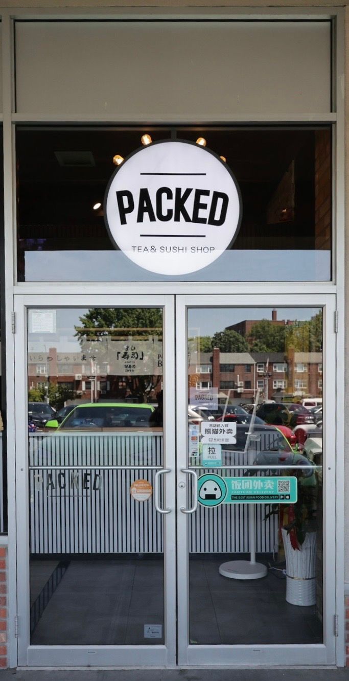 Storefront with a round “PACKED” sign above glass double doors, reflecting parked cars outside.