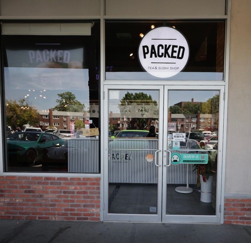 Storefront with a circular “PACKED” sign, glass doors, and brick facade reflected from the parking lot.