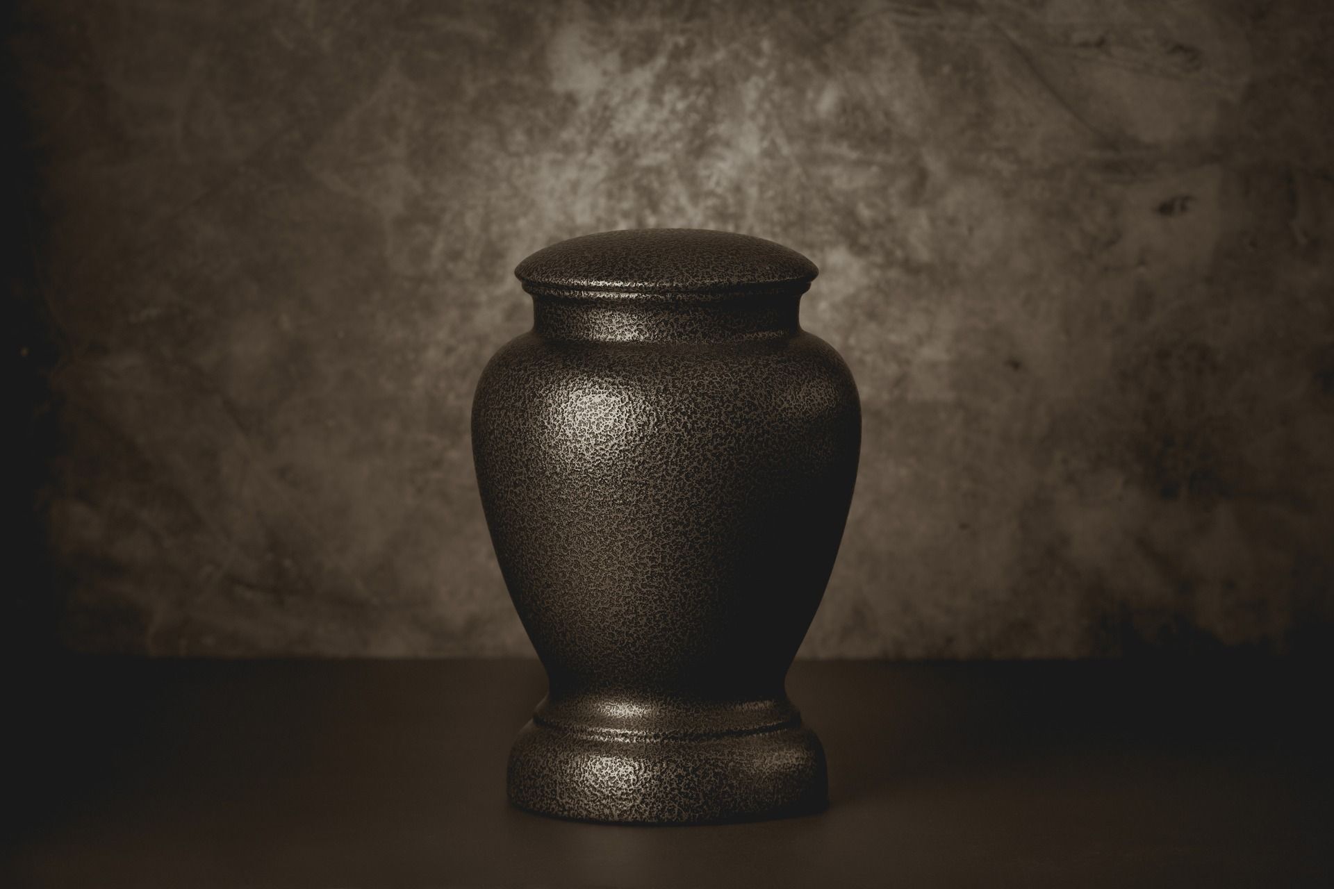 A textured, dark brown funeral urn centered on a dark surface against a mottled, neutral-toned background.