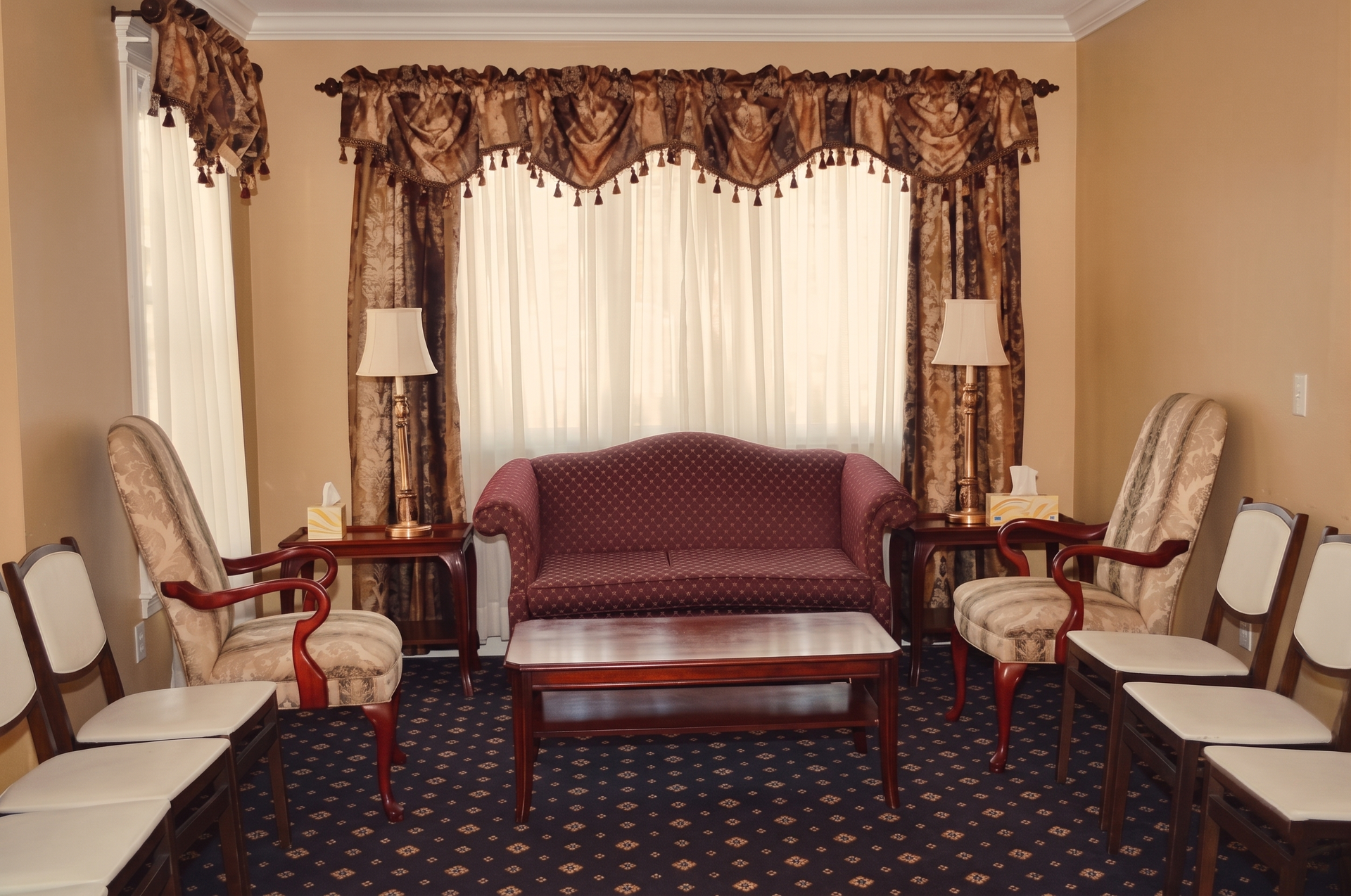 A formal waiting room with a maroon loveseat, patterned armchairs, wooden tables with lamps, and rows of chairs on carpet.
