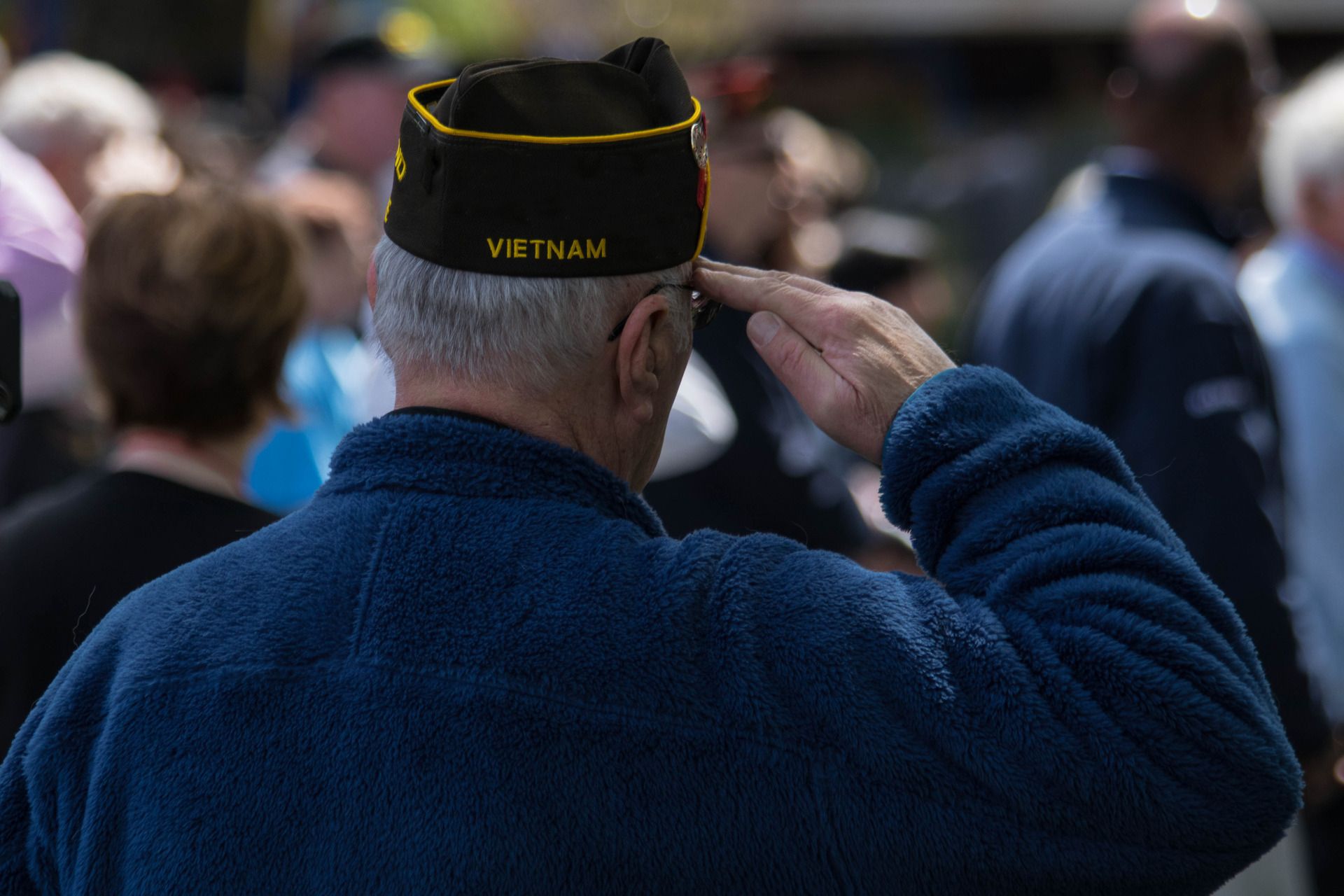 A veteran wearing a Vietnam cap salutes while standing in a crowd during an outdoor event.