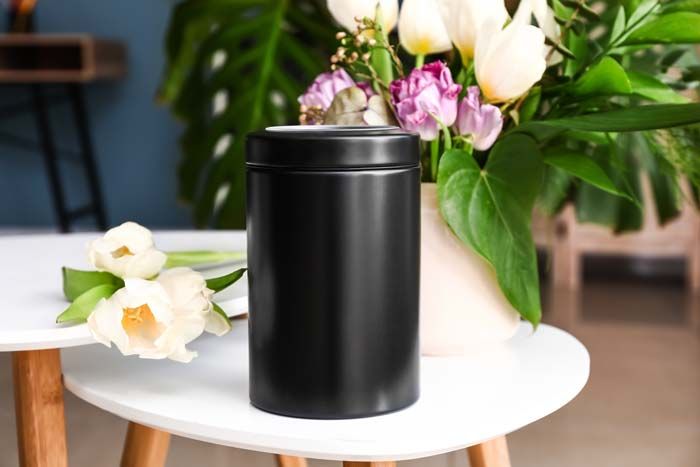 A black cylindrical cremation urn sits on a white table next to a bouquet of flowers.