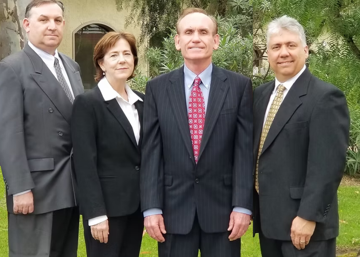 Four people in business attire standing outdoors; two men on each side, woman in the middle.