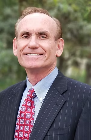 Man in suit smiles, red patterned tie, blurred outdoor background.