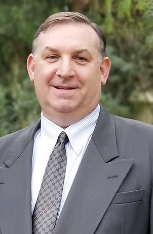 Man in a gray suit and patterned tie, smiling. Outdoor background.
