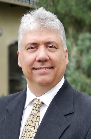 Man in a suit smiles, outdoors. Light skin, gray hair, patterned tie.