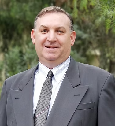Man in a gray suit and tie, smiling, outdoors with trees in the background.