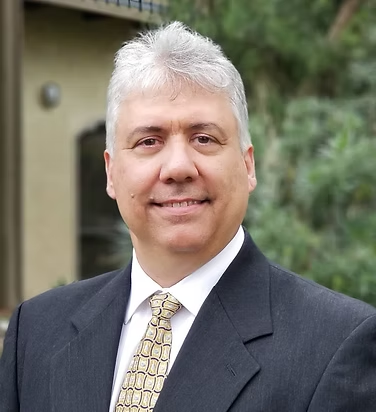 Man in a suit smiles, outdoors. Gray hair, patterned tie, building in background.