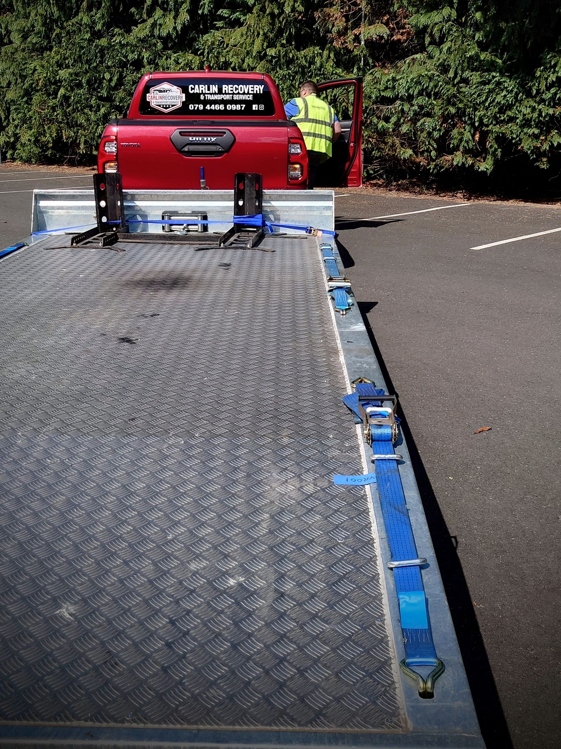 A red truck is towing a vehicle recovery trailer with winch and motorcycle stands in a parking lot