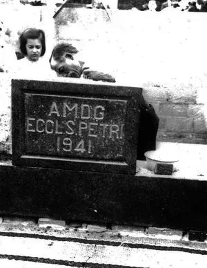 Black-and-white photo of a grave marker reading “AMDG ECCLES PETER 1941,” with two children beside it.