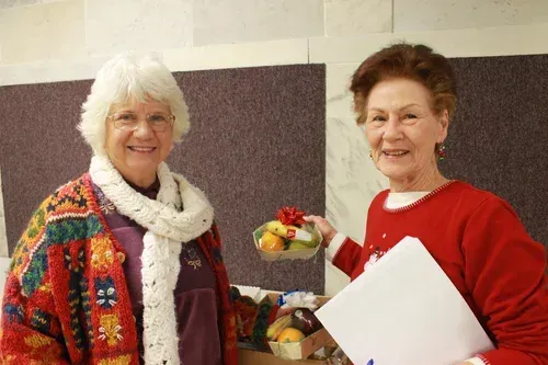 Two women smiling indoors, one holding a bowl of fruit and the other holding papers.