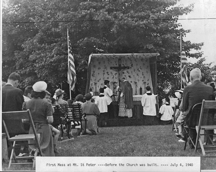 Outdoor church service with altar and cross, congregation seated on grass beneath trees, American flag nearby