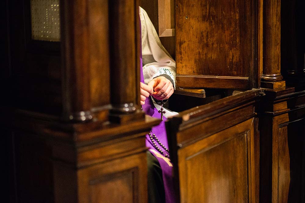 Person seated in a wooden confessional, hands clasped, wearing a purple garment.