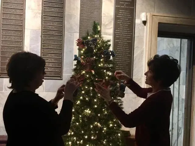 Two people decorating a lit Christmas tree indoors, seen in silhouette.