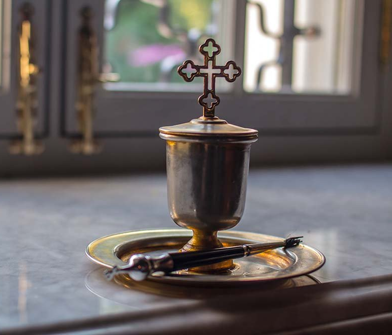 A silver chalice with a cross lid on a plate, lit by sunlight in a church-like setting.