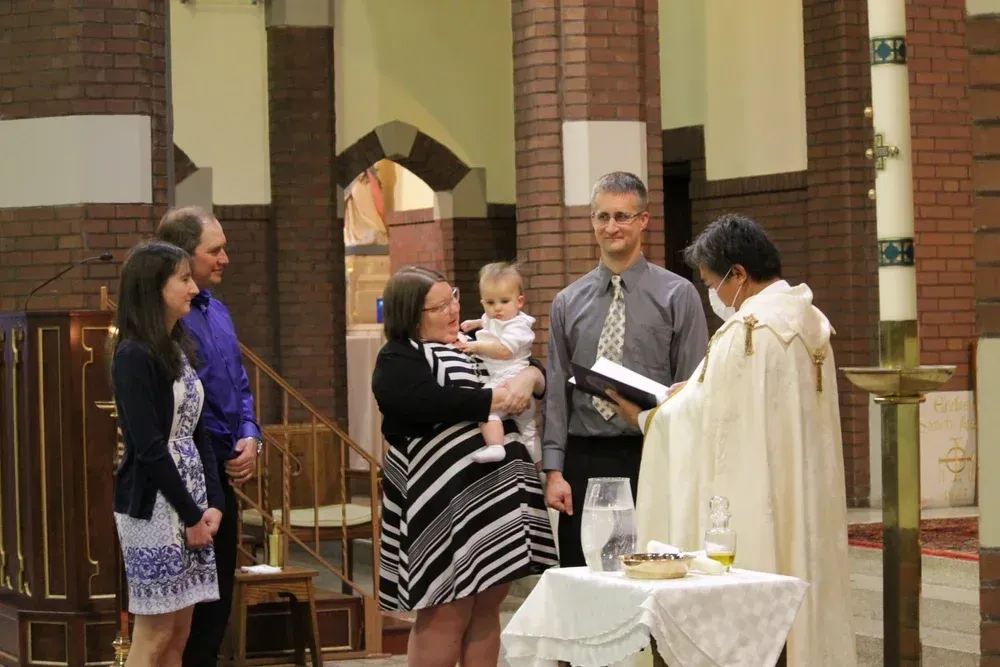 Priest blessing a baby during a church ceremony, with family gathered nearby.