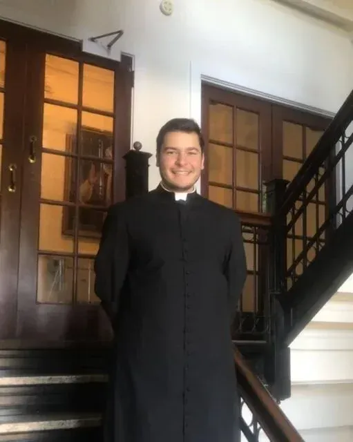 Person in a black clerical robe standing in a staircase landing, smiling.
