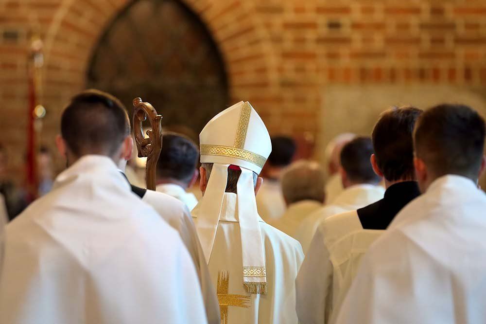 Clergy in white robes gathered in a church, with a bishop in a white mitre facing forward.
