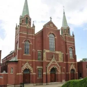 Red brick church with twin green spires and arched entrance against a cloudy sky