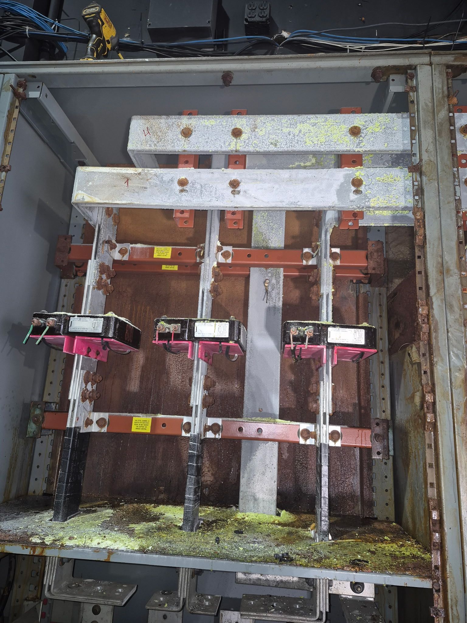 Internal view of an industrial electrical cabinet with corroded busbars, loose debris, and three electrical switch units.