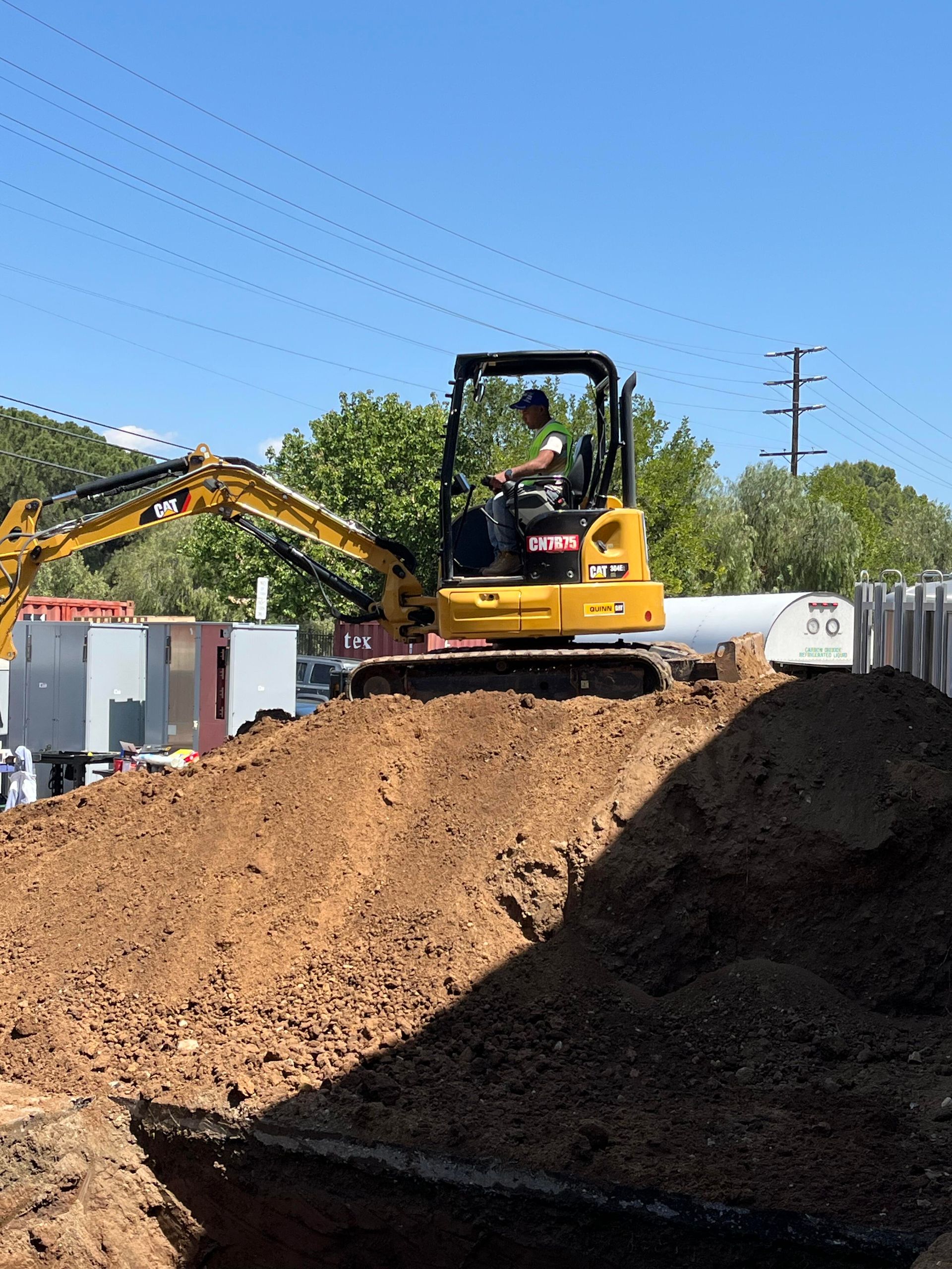A yellow mini excavator sits atop a large pile of dirt at a sunny construction site.