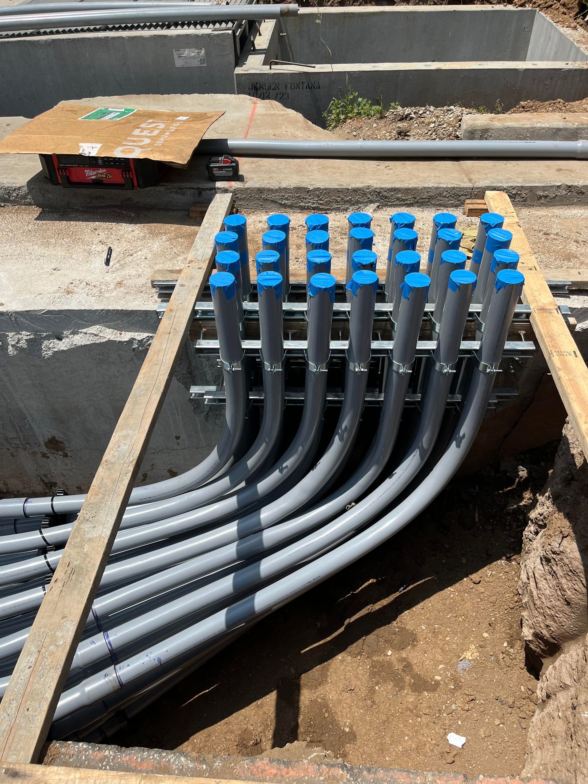 A series of gray electrical conduits with blue caps emerging from a trench and secured in a row on a metal rack.