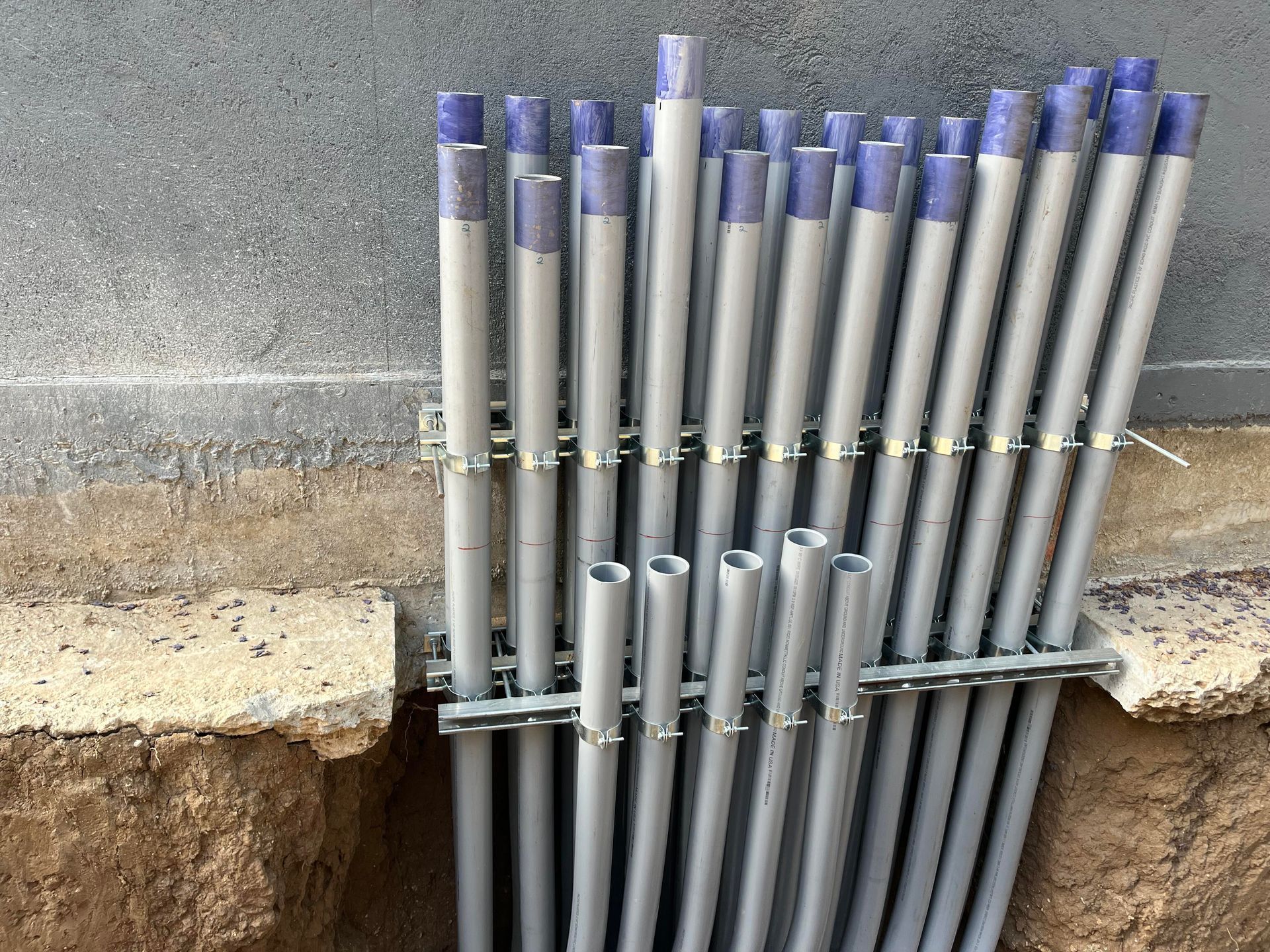 A cluster of gray electrical conduits held by metal clamps against a concrete wall foundation above a trench.
