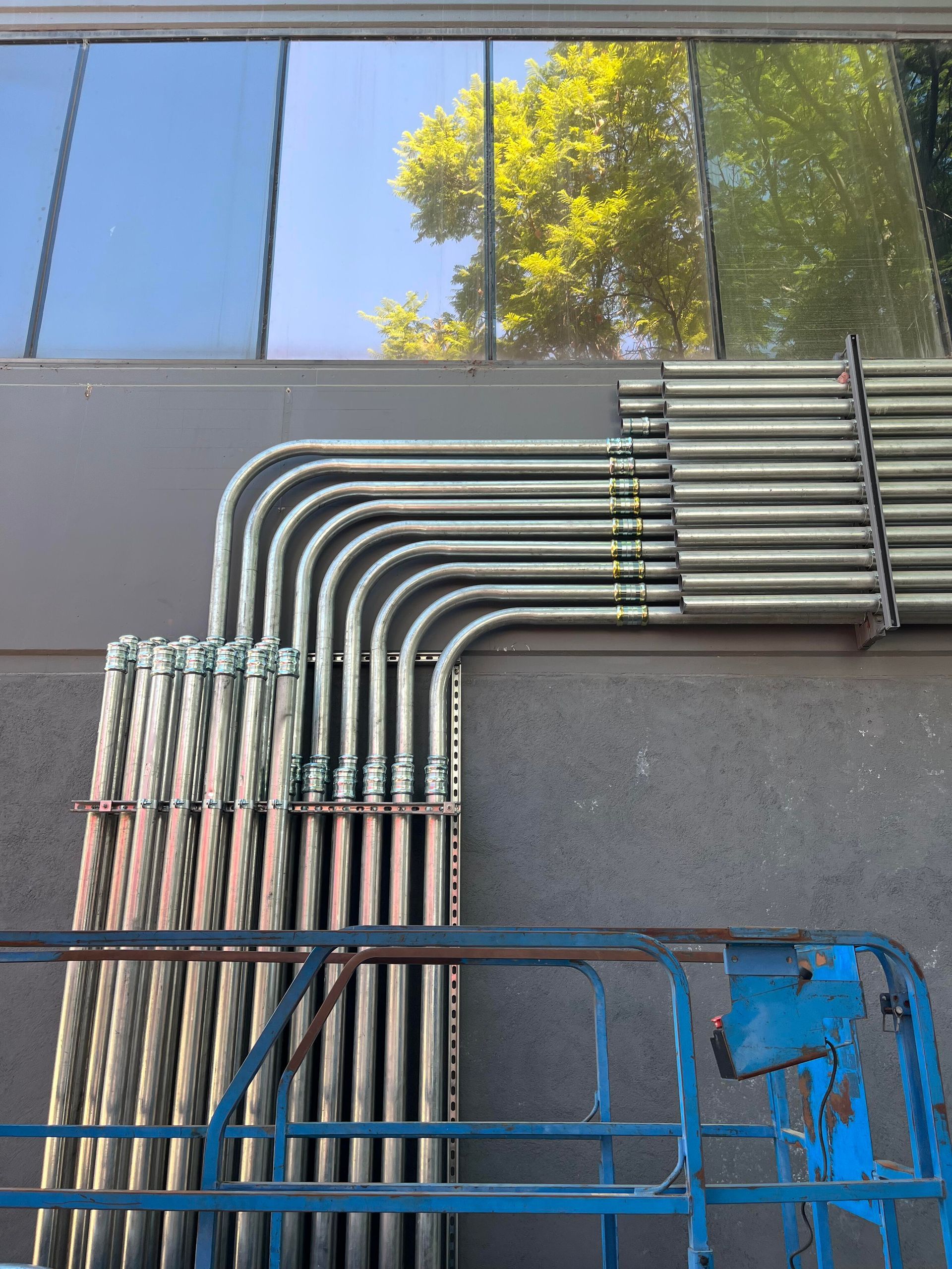 Rows of silver conduit pipes installed on a gray exterior wall, forming precise 90-degree bends.