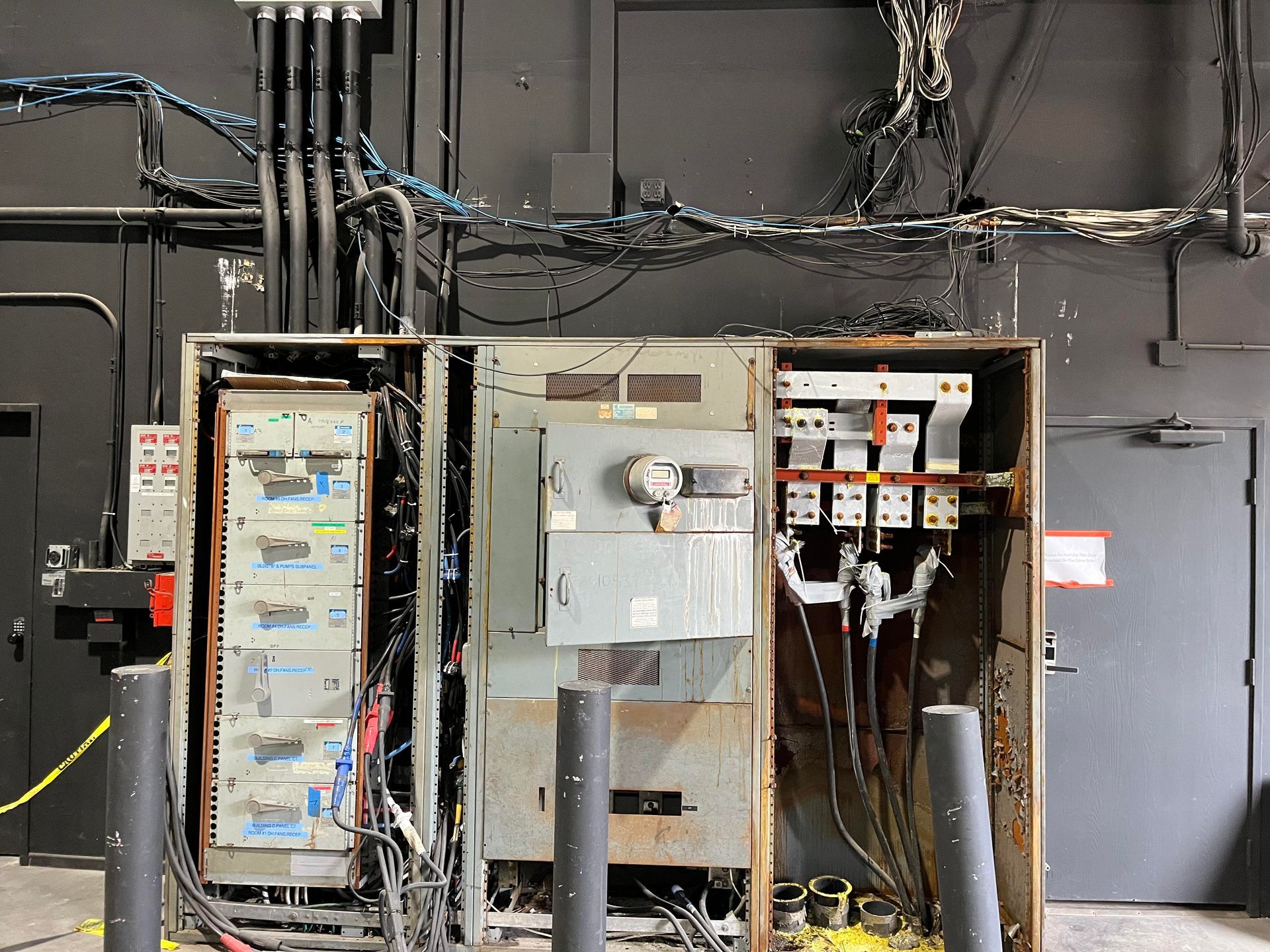 Industrial electrical equipment with exposed wiring, grey metal cabinets, and overhead conduits against a dark wall.
