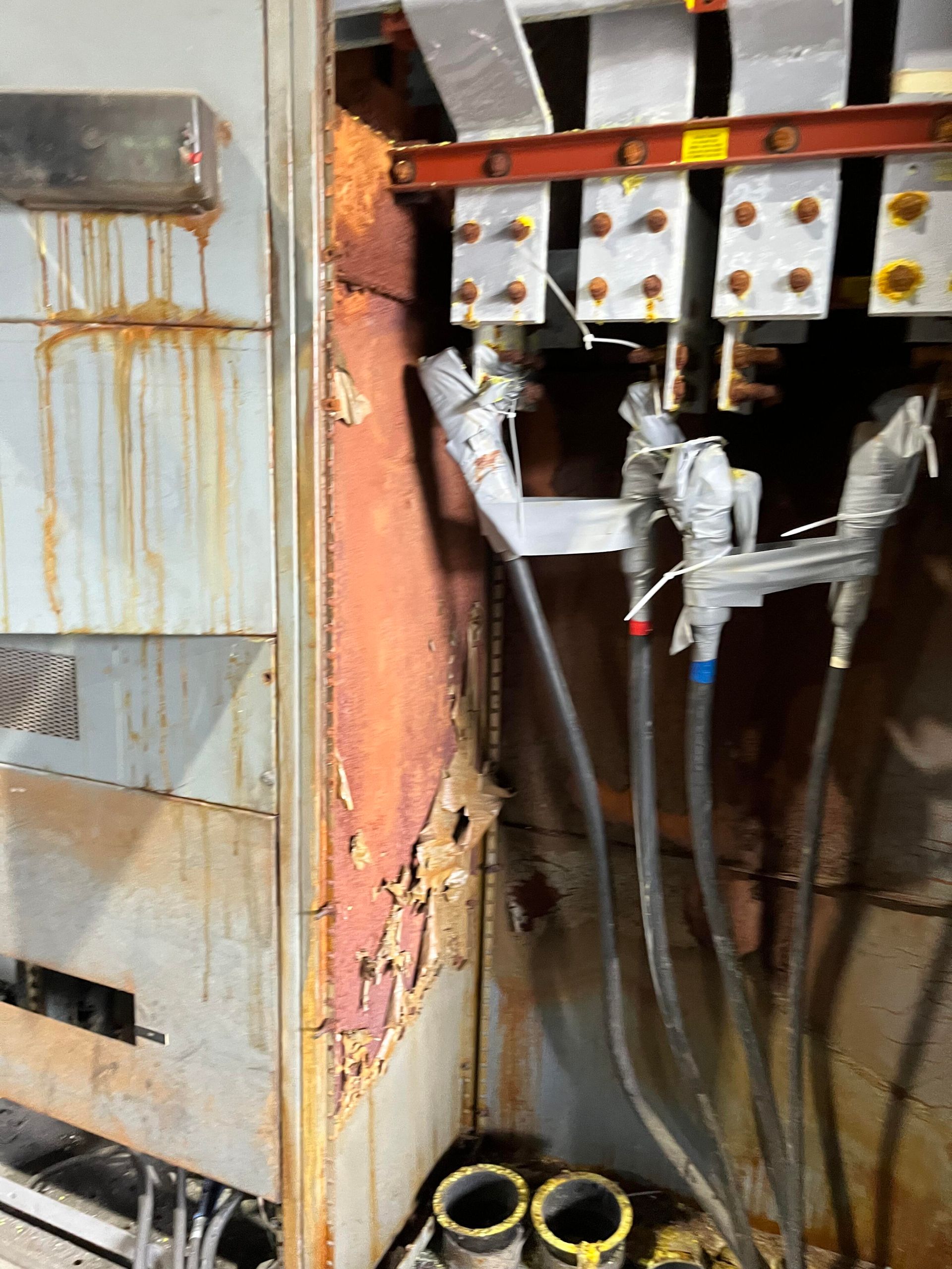 Electrical panel with heavy corrosion and water staining on the metal cabinet and frayed, taped wiring connections.