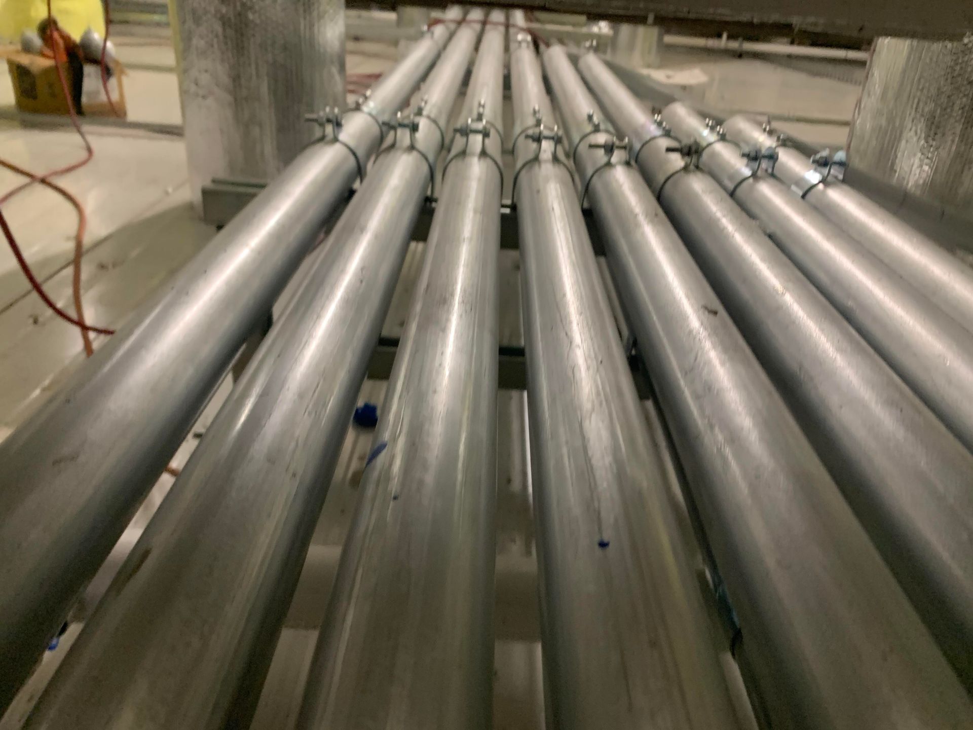 Multiple silver industrial pipes with metal clamps, arranged in parallel lines extending into an indoor facility.