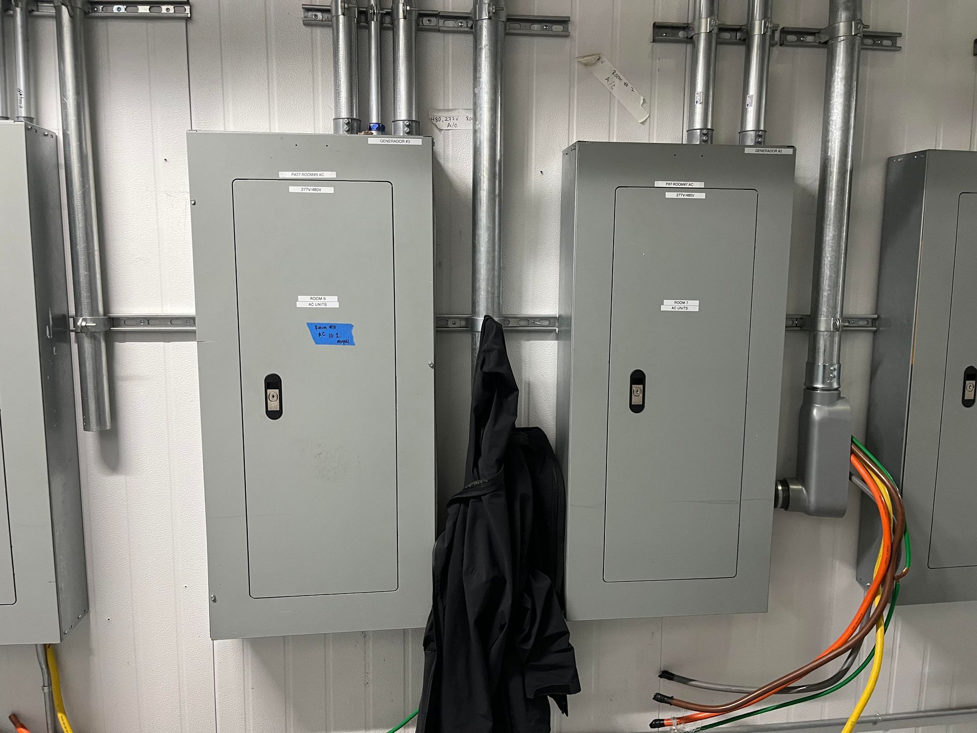 Electrical breaker panels mounted on a wall with metal conduits and exposed, colorful wiring hanging from the right side.