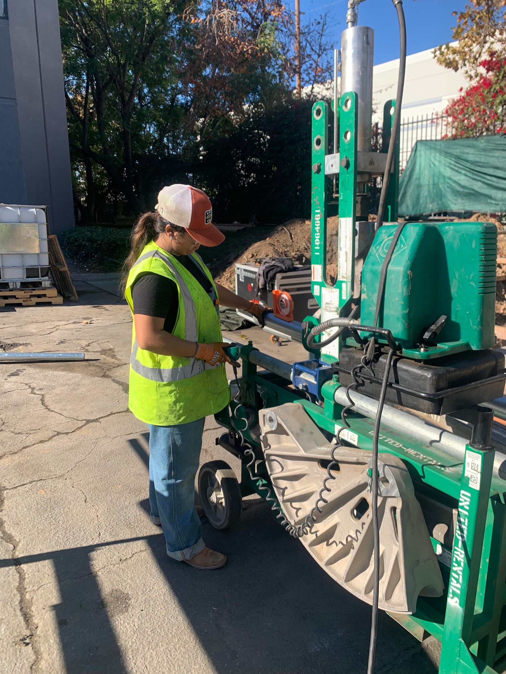 A person wearing a high-visibility vest and a cap operates a green machine outdoors in a paved work area.