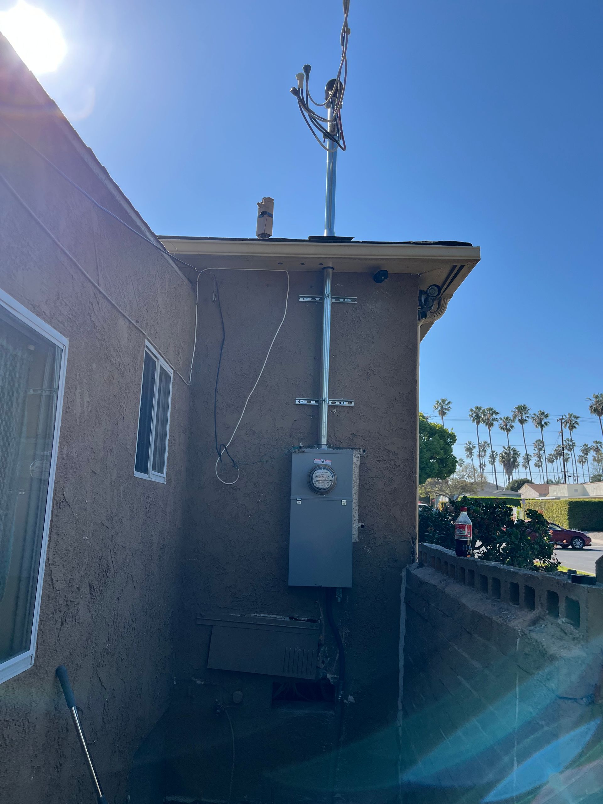 Exterior view of a home showing an electrical meter box mounted on a stucco wall with a conduit extending to the roof.