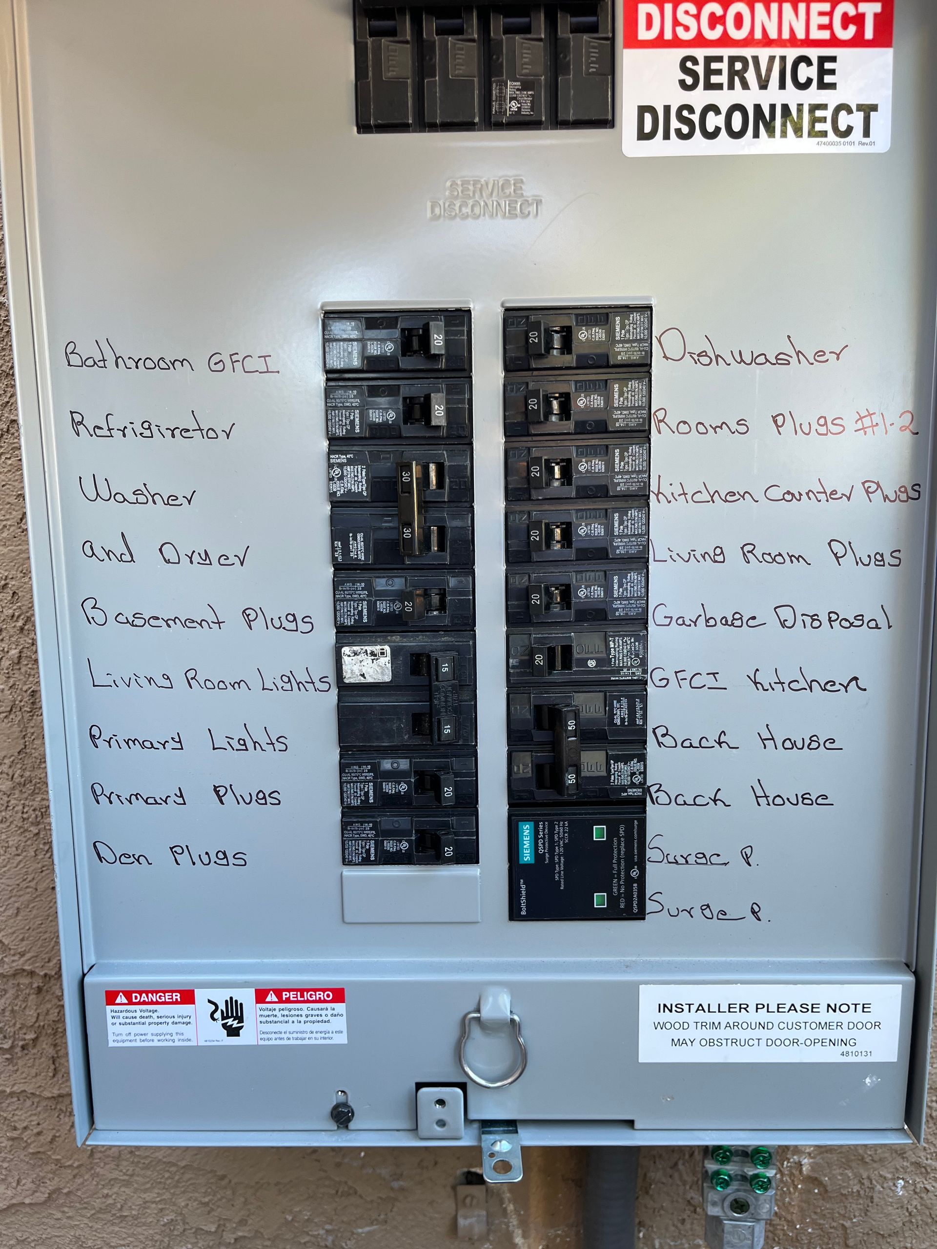 A gray electrical breaker panel mounted on a textured wall, with labels identifying various home circuits.