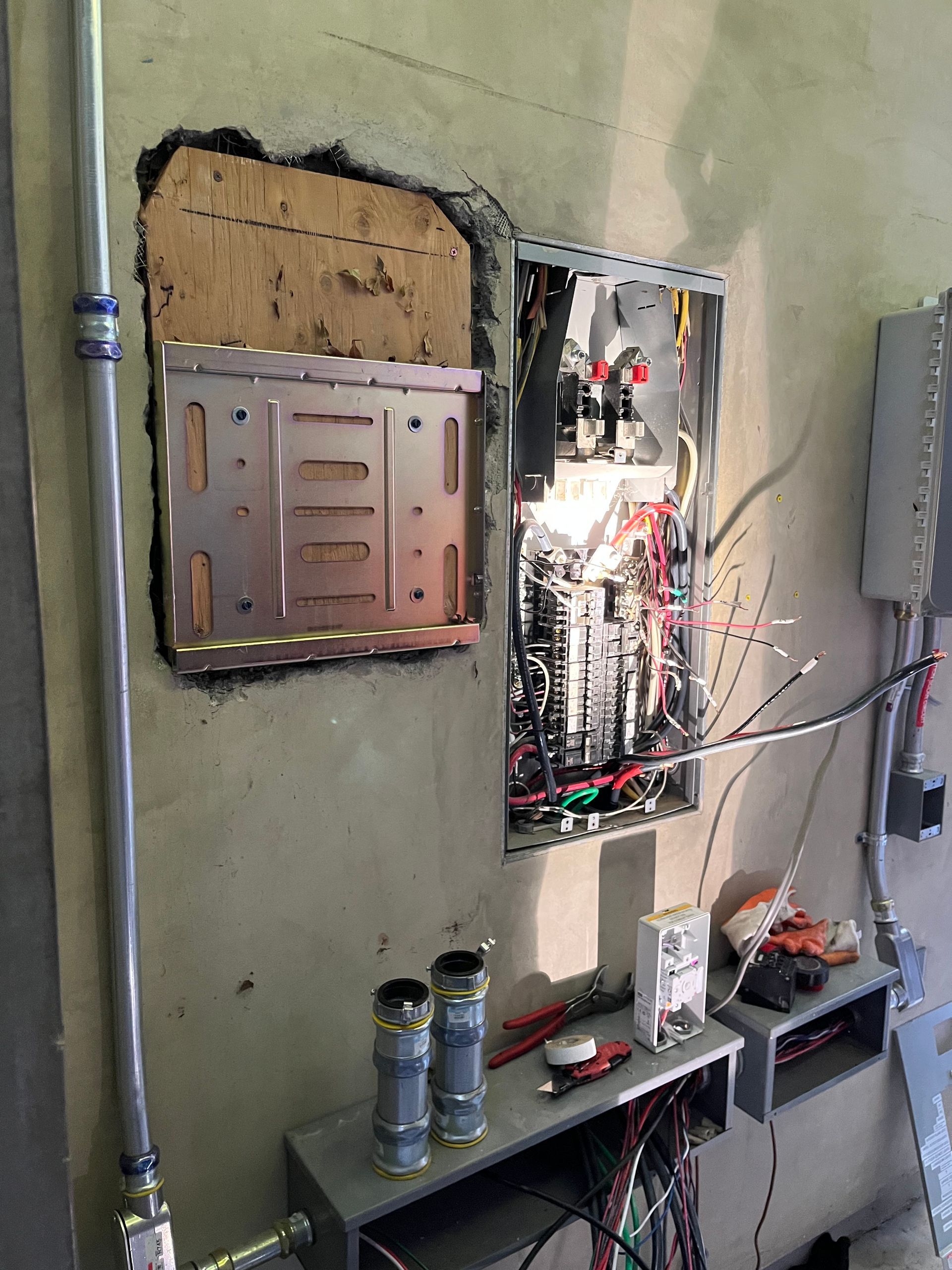 Electrical work in progress on a concrete wall featuring an open circuit breaker panel and a mounting bracket nearby.