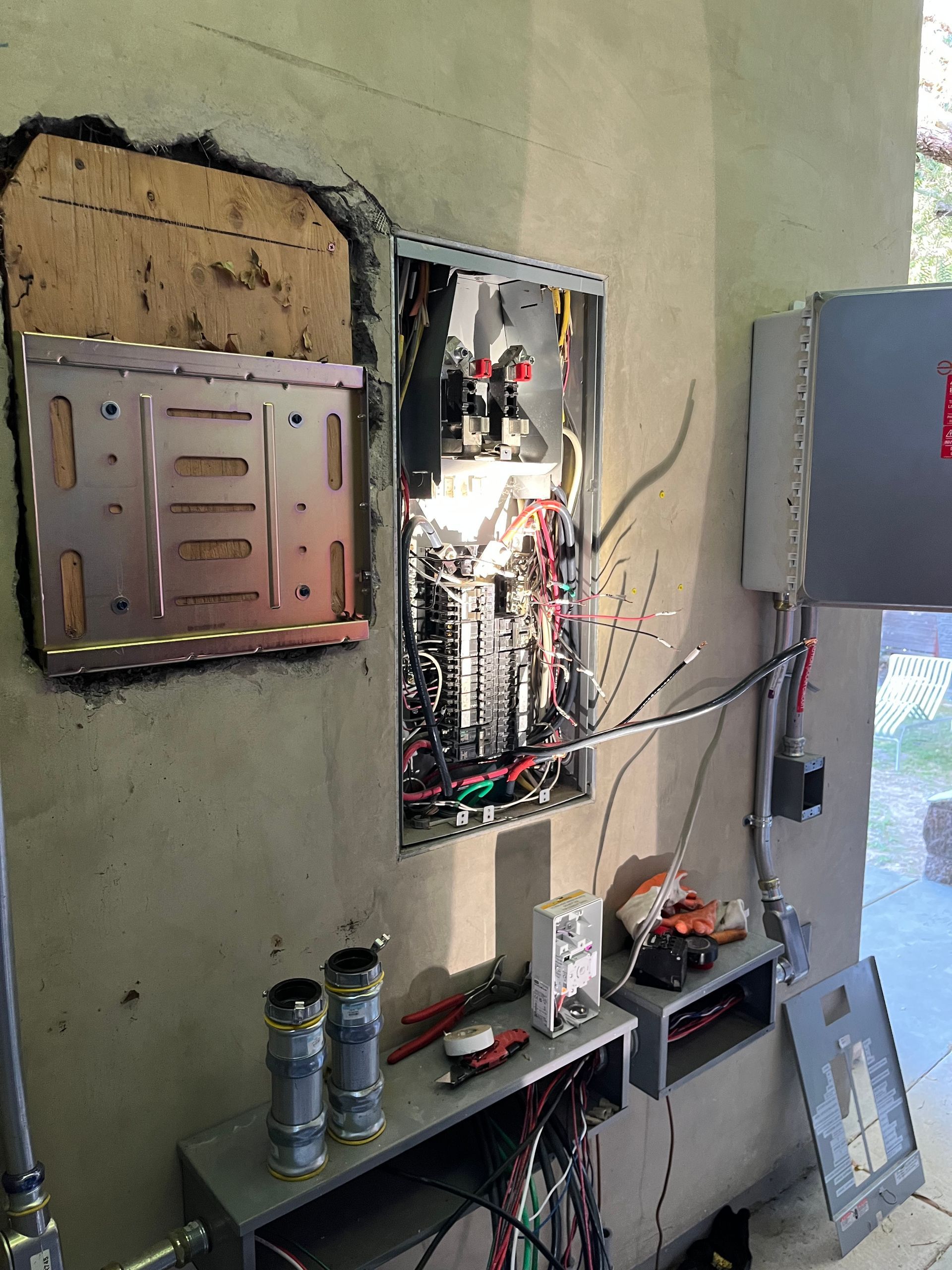 An open electrical breaker panel mounted on a concrete wall, with exposed wires and wall-mounted metal hardware nearby.