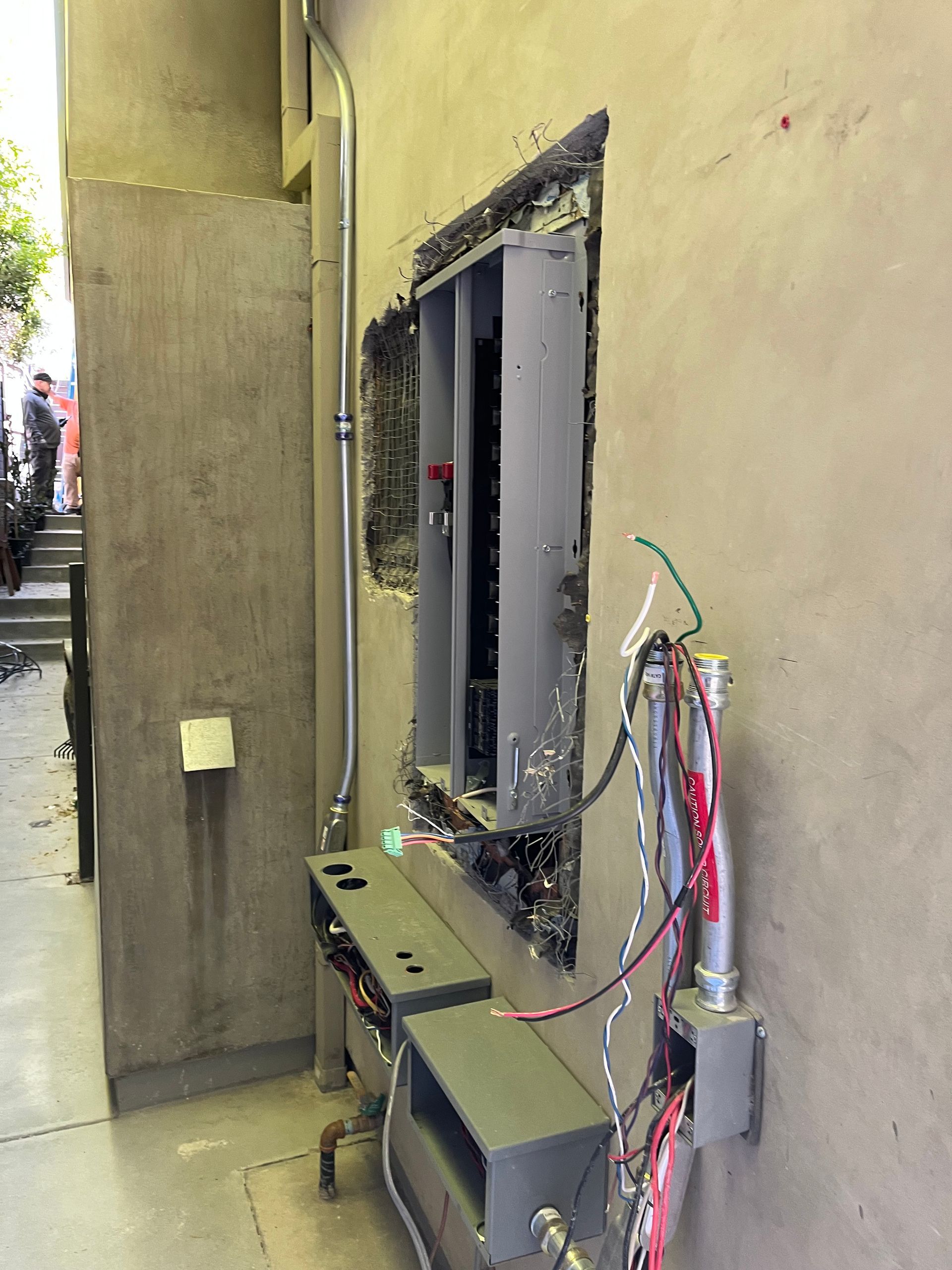 An electrical panel housing sits partially installed on a beige exterior wall, surrounded by exposed wiring and conduits.