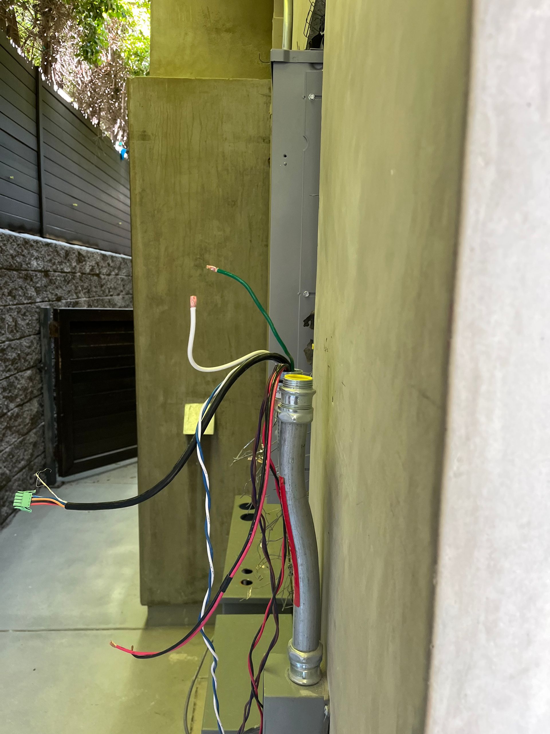 Exposed electrical wires extending from a metal conduit on an exterior stucco wall near a fence.