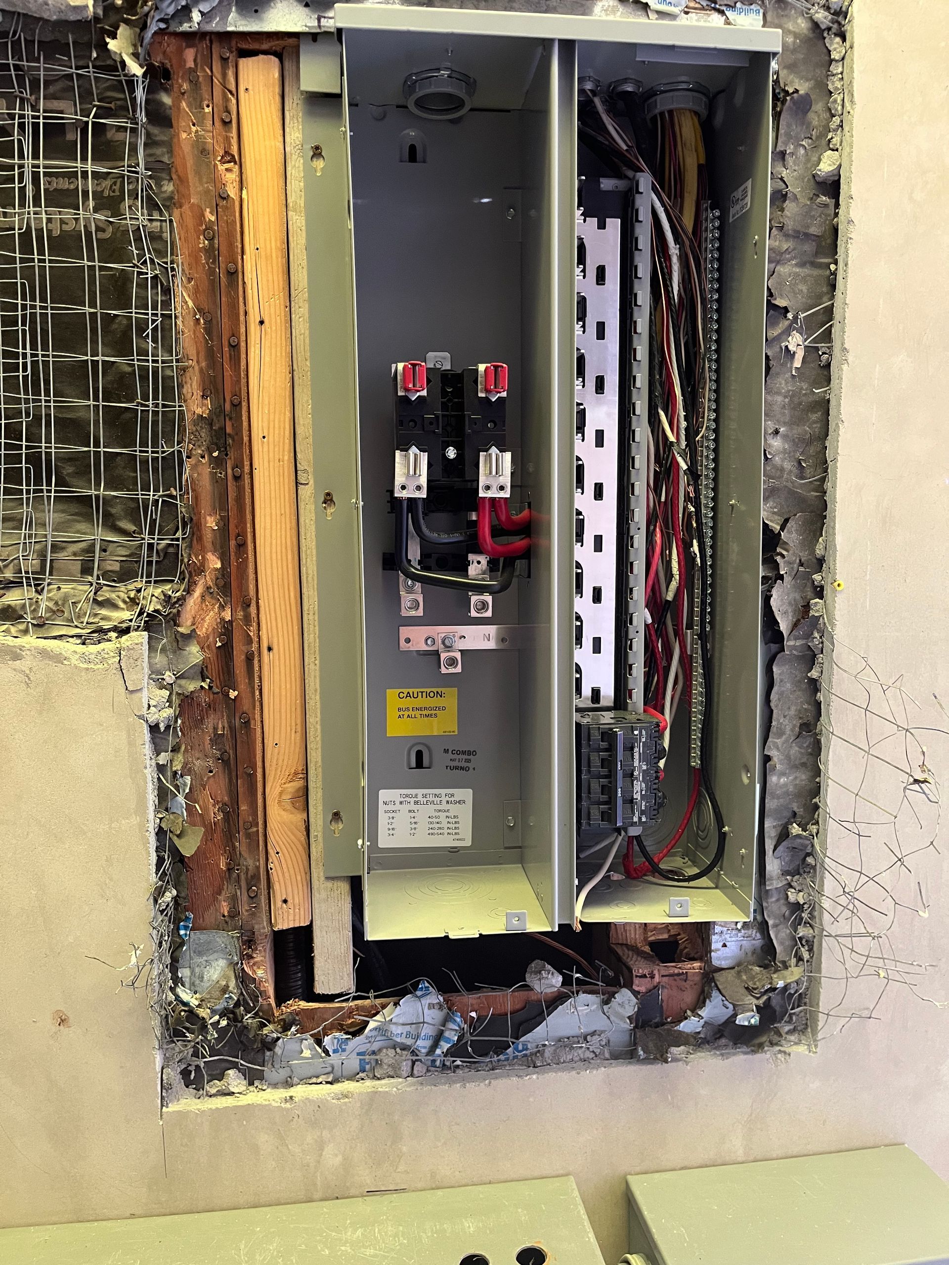 An electrical panel box set into a wall, with wires visible and drywall removed from around the housing.