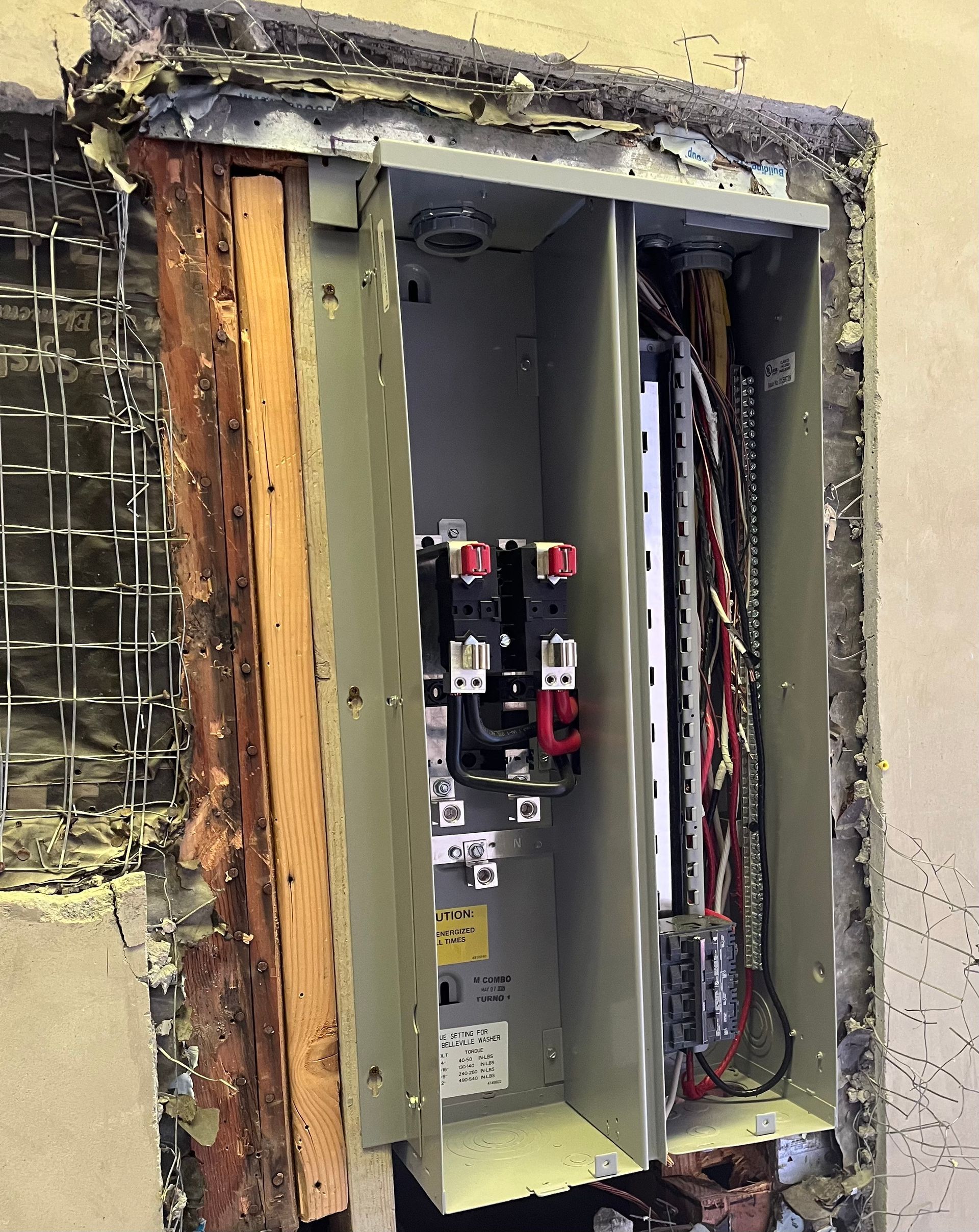 An open electrical panel recessed in a wall, revealing interior wiring, circuit components, and insulation.