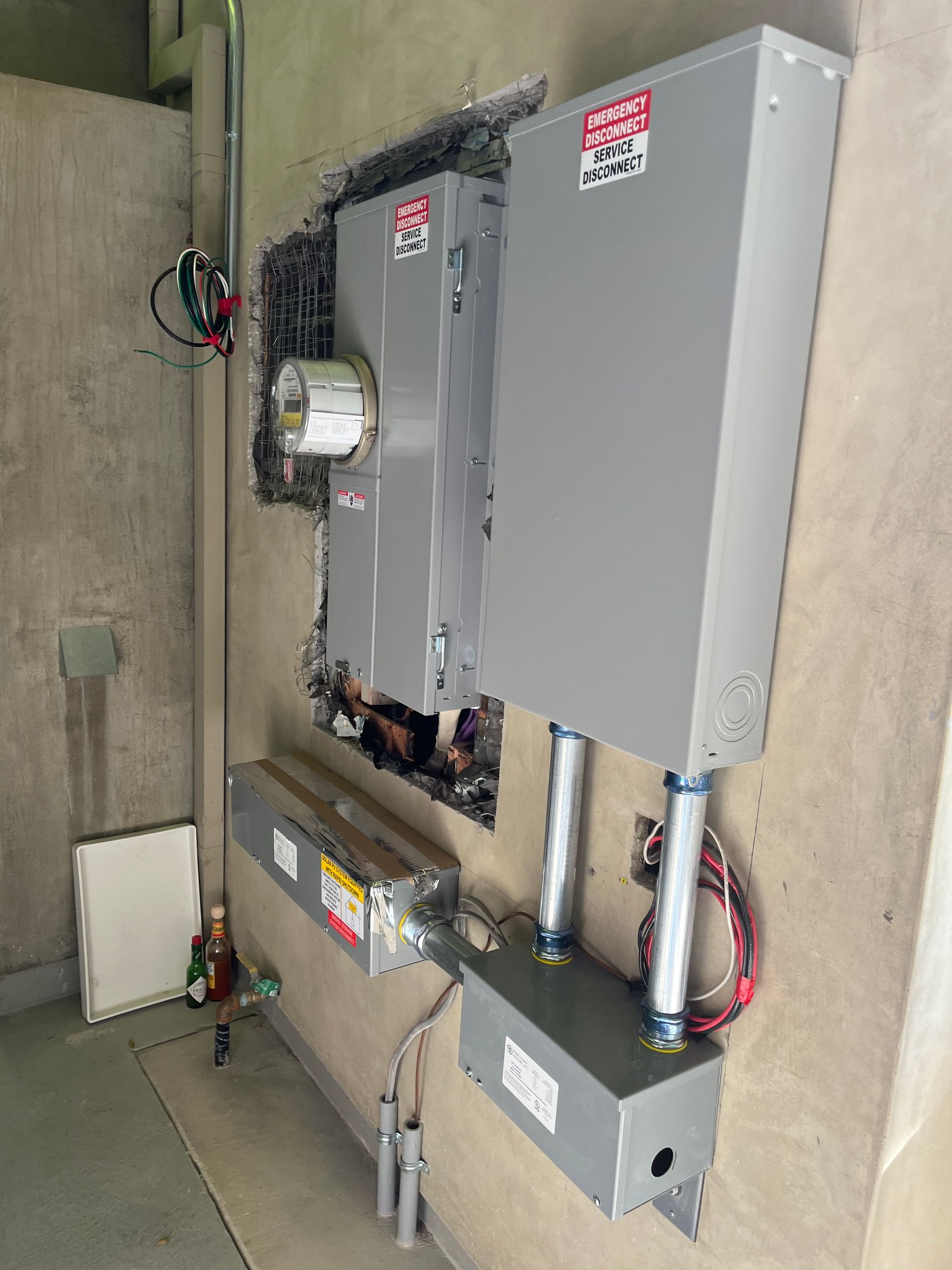 A residential electrical meter base and service disconnect panel mounted on a gray wall with associated conduit and wiring.